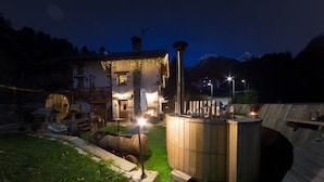 A rustic wooden building with a stone chimney, a wooden hot tub, and a well-lit outdoor area at night. - La Maison d'Antan (Bionaz)