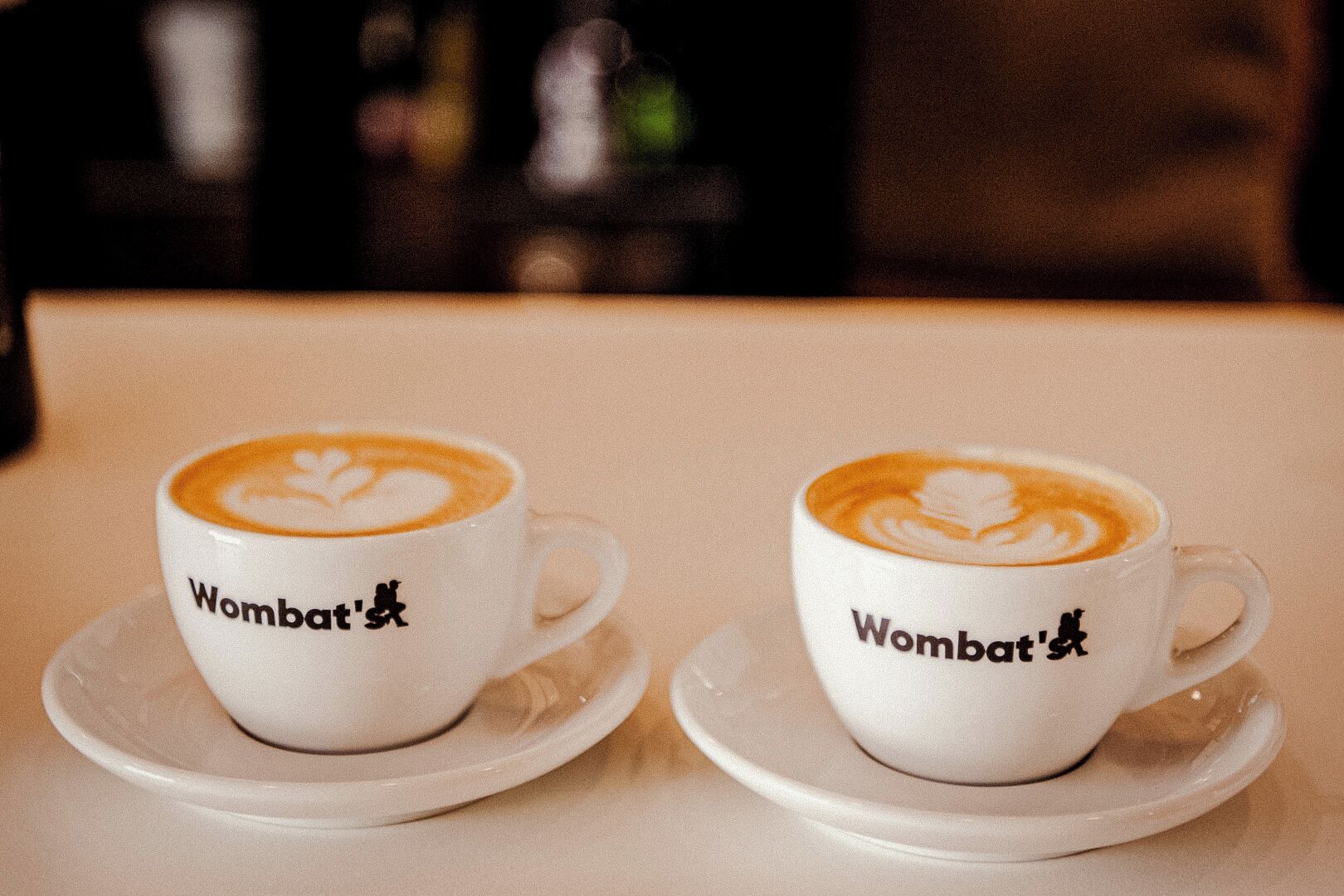 Dos tazas blancas con arte de leche y el texto 'Wombat's' en un plato.