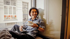 Una persona sentada junto a una ventana, con una taza, una bolsa y una cámara.