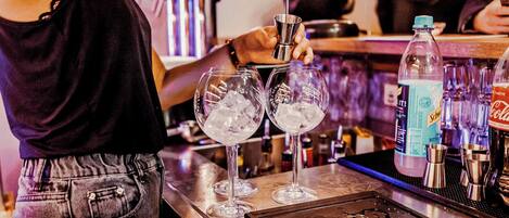 Un bar con un bartender preparando bebidas, vasos con hielo, y diferentes botellas y utensilios de bar.