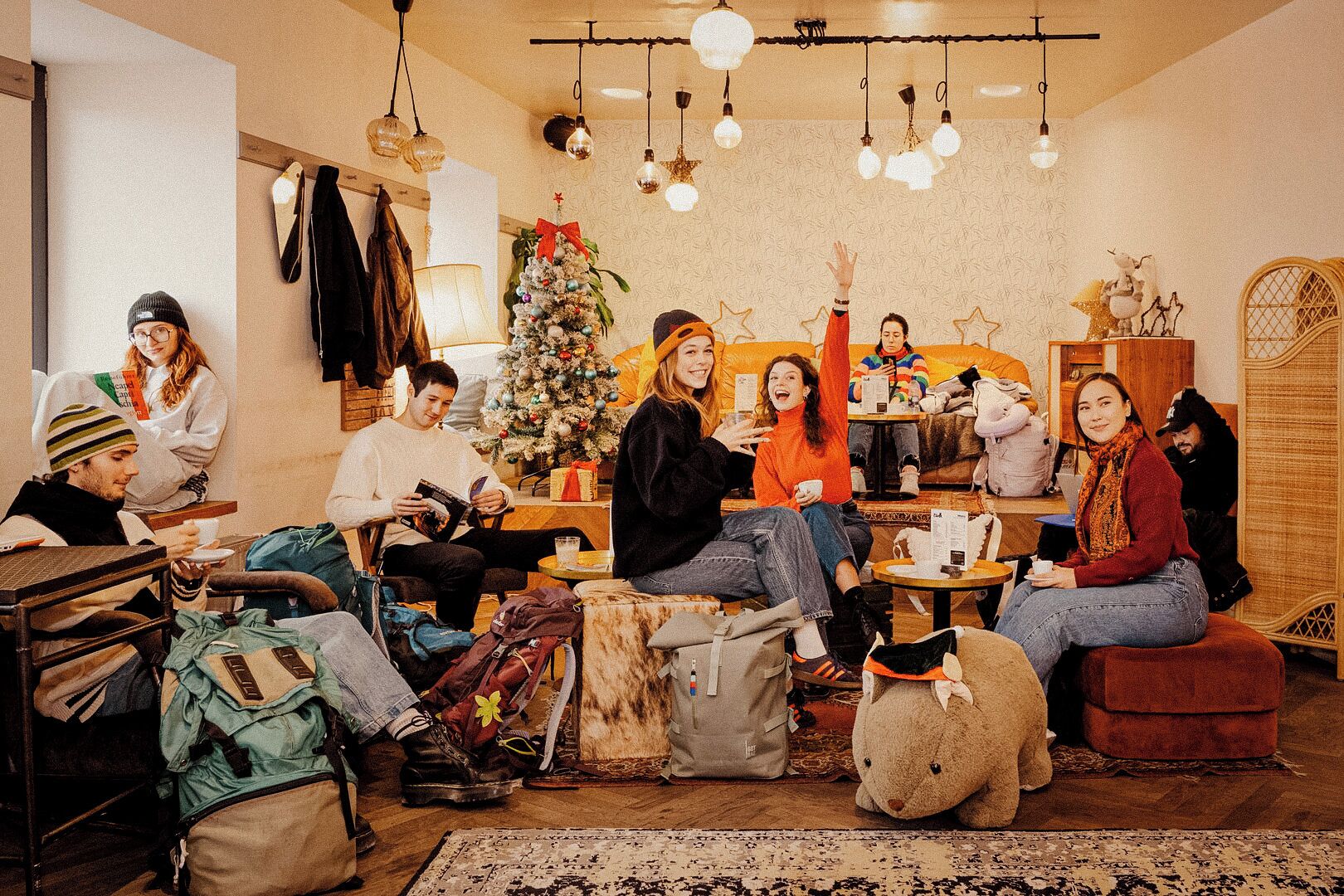 A cozy indoor setting with a group of people gathered around a table, a Christmas tree, and various seating arrangements.
