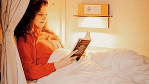 Un lit simple avec une tête de lit, une table de chevet avec une lampe et une étagère fixée au mur, sur laquelle est posé un panneau.