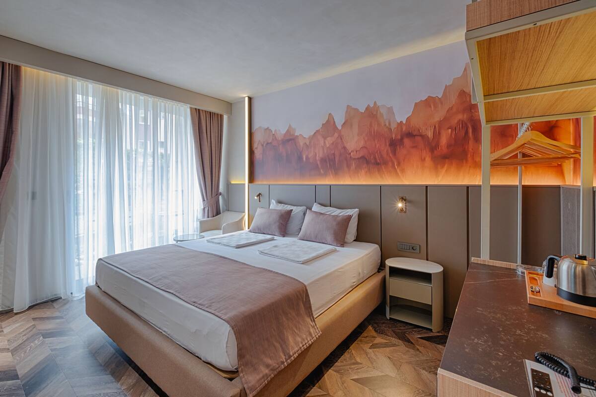 A modern hotel room with a large bed, a wooden desk, and a mountain landscape mural.