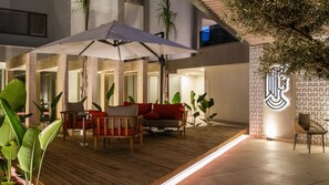 A modern outdoor seating area with wooden furniture and umbrellas, surrounded by large plants and a geometric wall design.