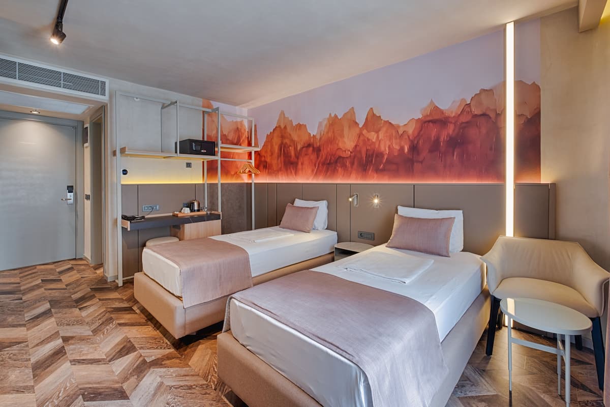 A hotel room with two beds, a desk, a chair, and a large mountain mural on the wall.