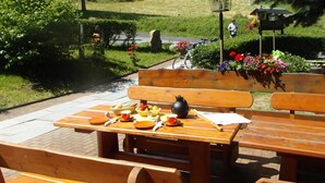 An outdoor dining area with wooden benches and tables, a picnic setup with food and drinks, and a scenic view of a hill with trees and a clear sky. - Sonnebergbaude Waltersdorf (Grossschoenau)