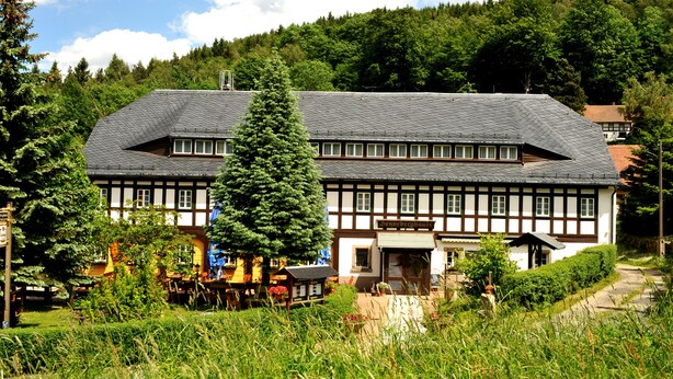 A traditional half-timbered building with a sloped roof, surrounded by lush greenery and a forested hillside. - Sonnebergbaude Waltersdorf (Grossschoenau)