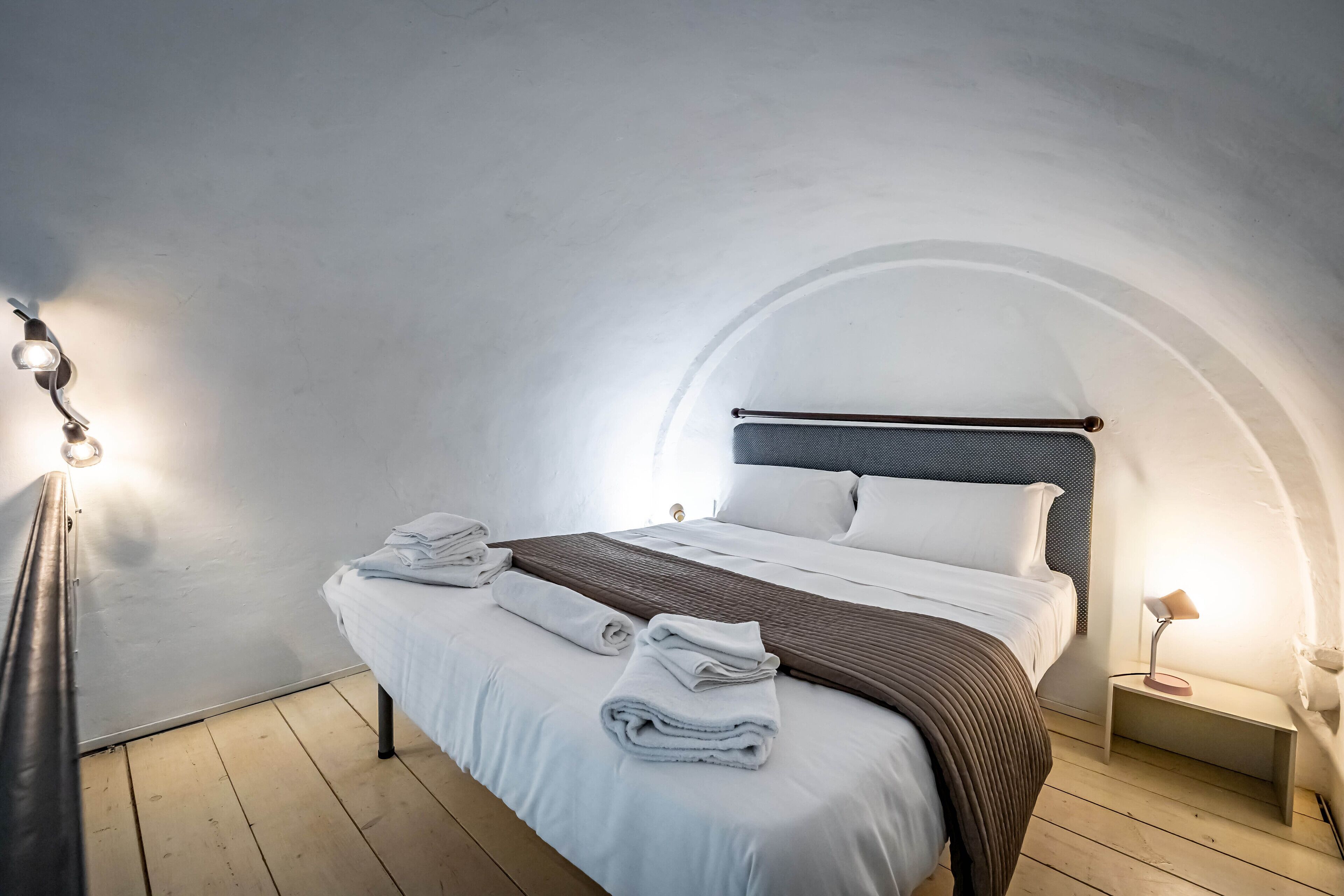 A neatly made bed with white linens and a brown throw blanket, a bedside lamp, and a small bedside table in a room with a curved ceiling.