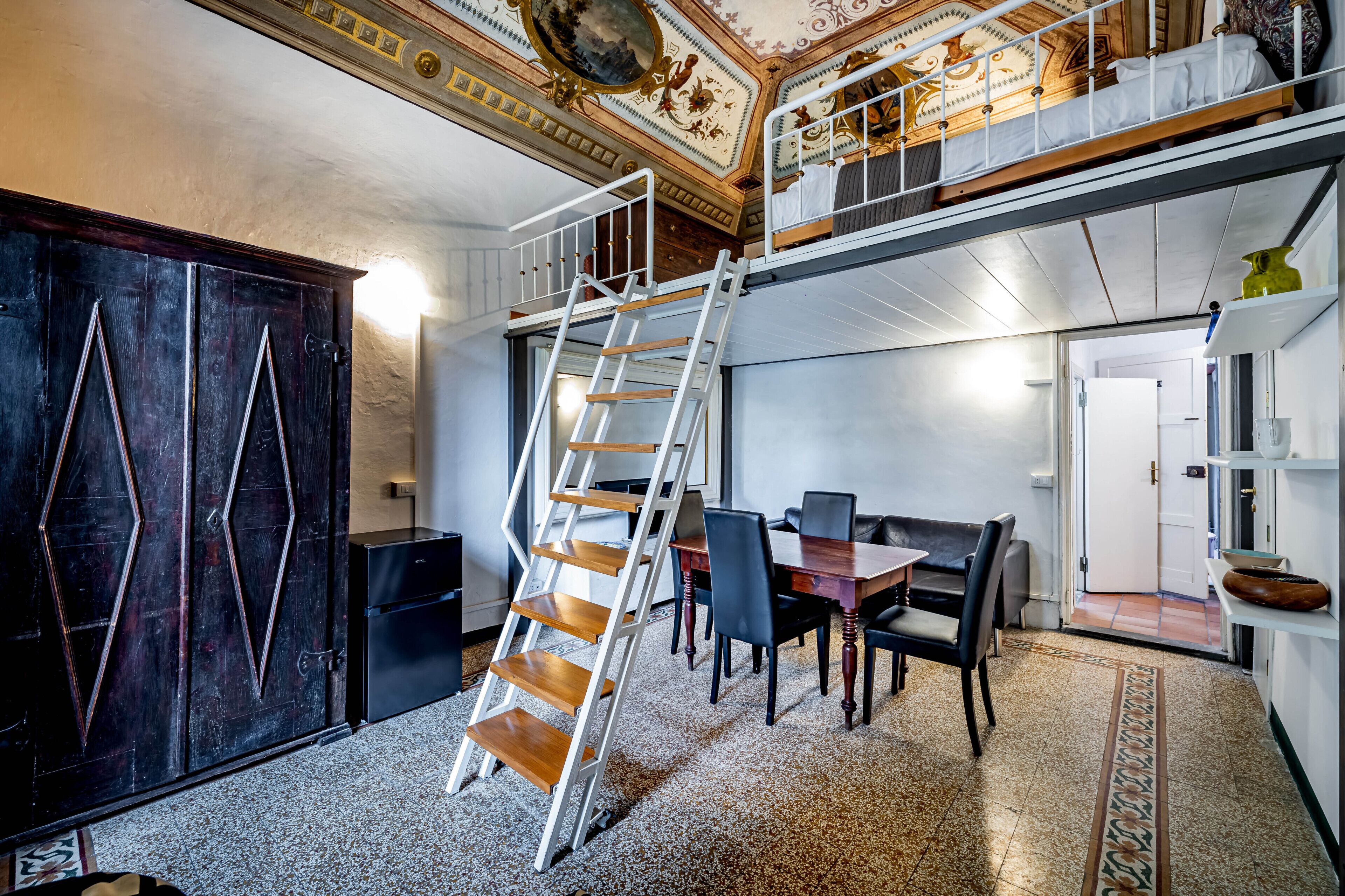 A room with a wooden staircase, a dining area with chairs and a table, a large dark wooden cabinet, and a ceiling with ornate designs.
