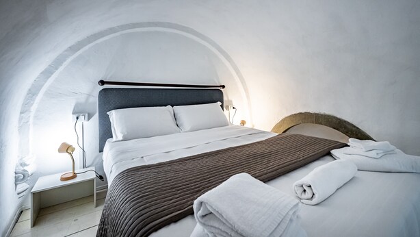 Een moderne slaapkamer met een bed, witte lakens, een bruine plaid en een nachtkastje met een lamp.