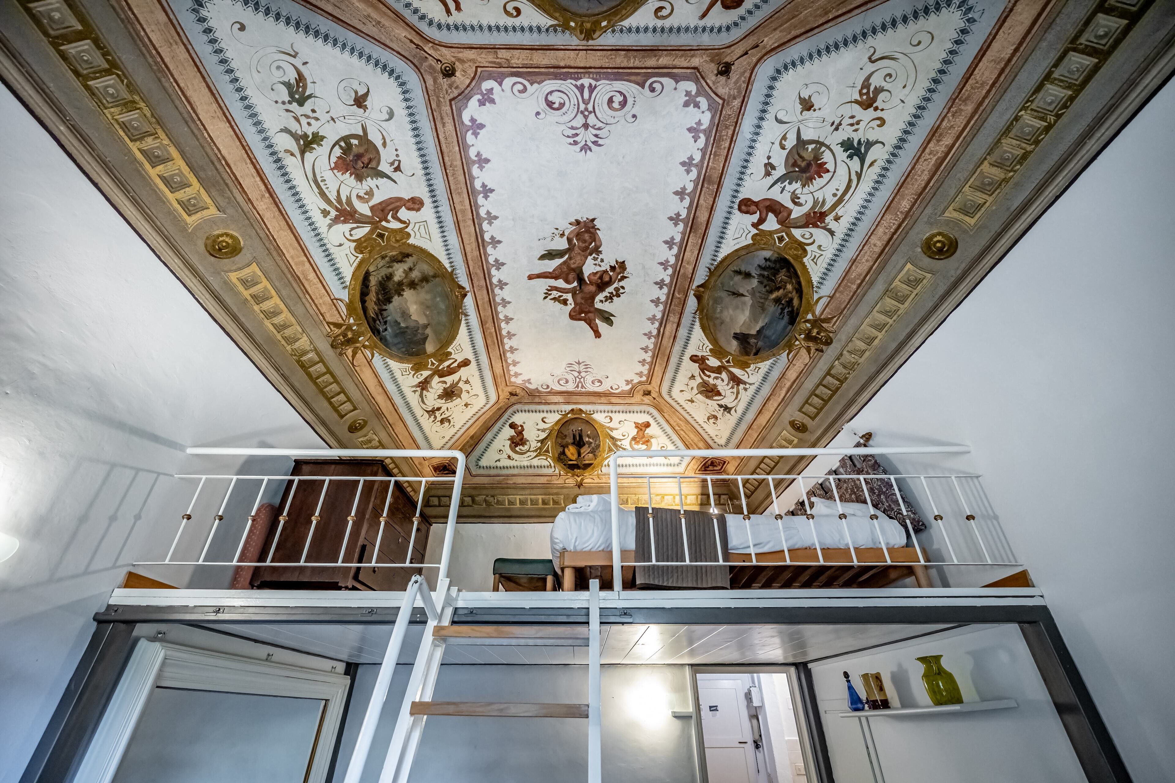 Et værelse med køjeseng, et lille skrivebord og et loft med detaljerede freskomalerier.