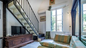 Een woonkamer met een trap, een televisie op een houten kast, een bank met gestreepte kussens, en een raam met gordijnen.