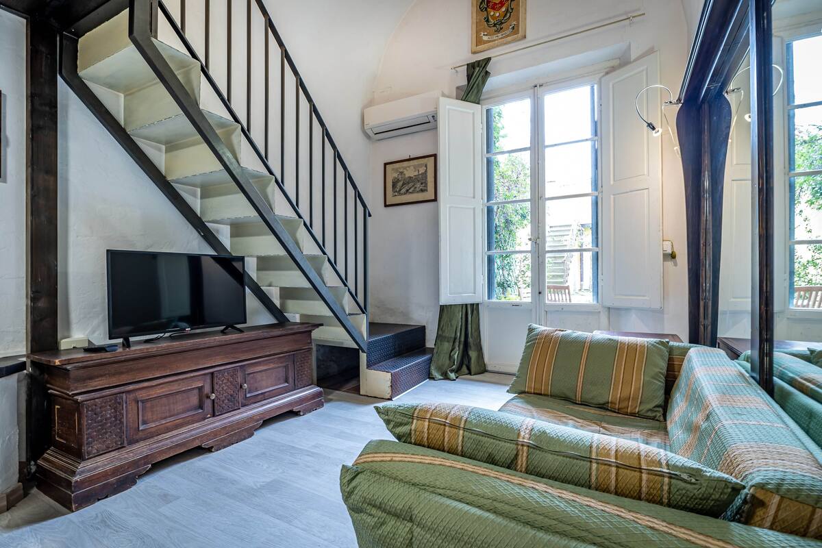A living room with a staircase, a television on a wooden cabinet, a sofa with striped cushions, and a window with curtains.