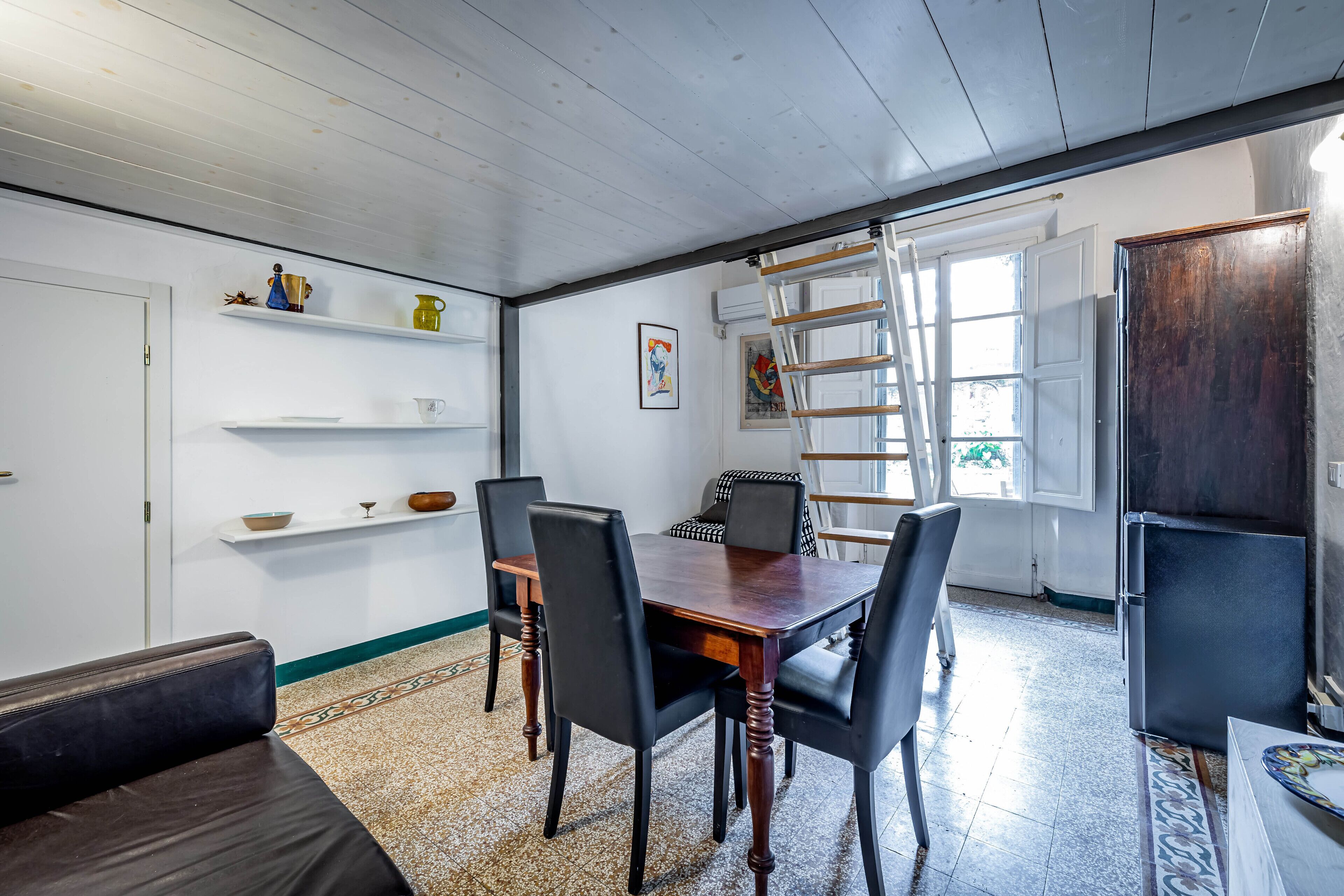A dining area with a wooden table and chairs, a sofa, and a ladder leaning against a door.