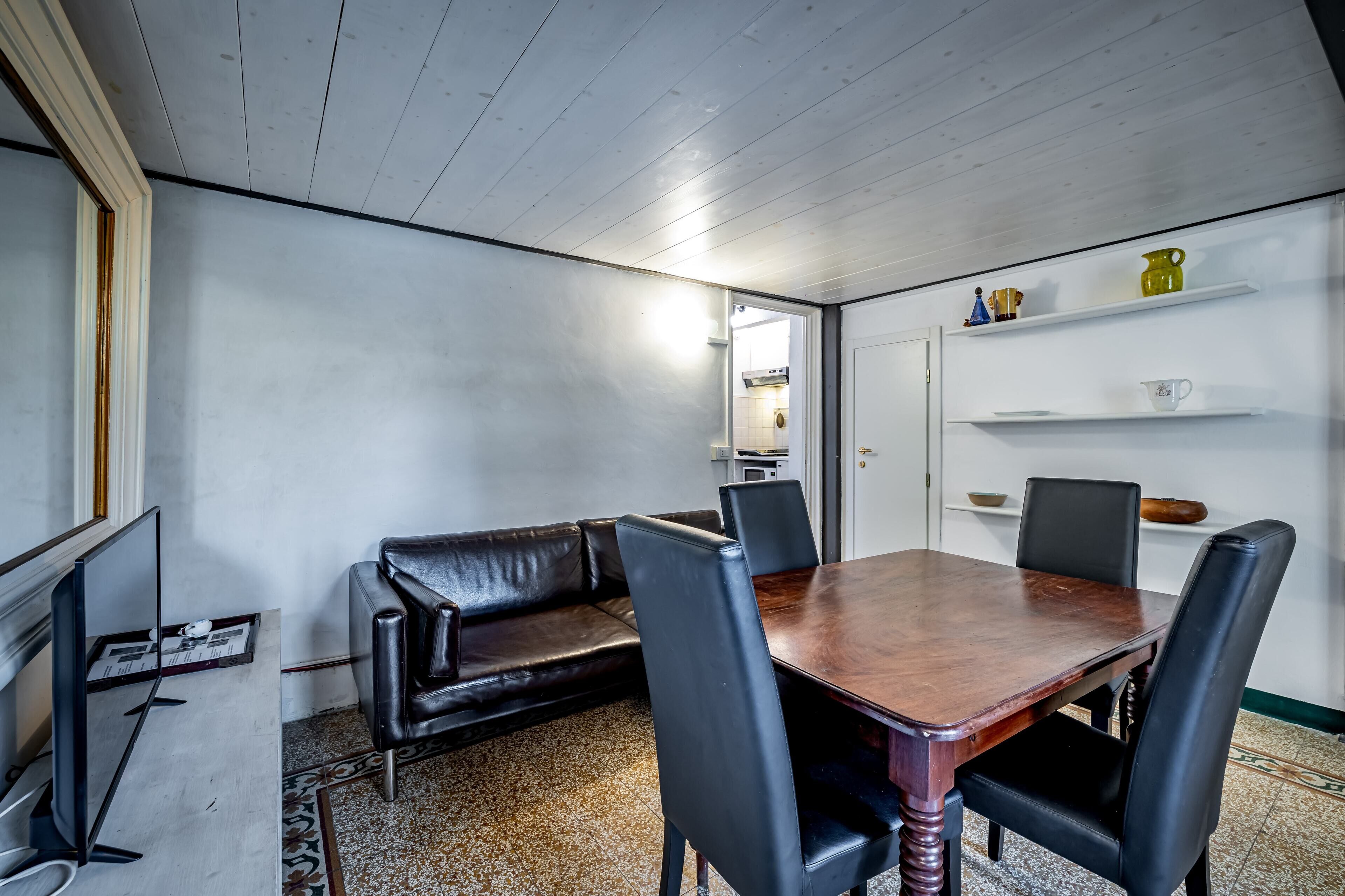 A dining area with a wooden table, black leather chairs, a black leather sofa, and a television on a stand.
