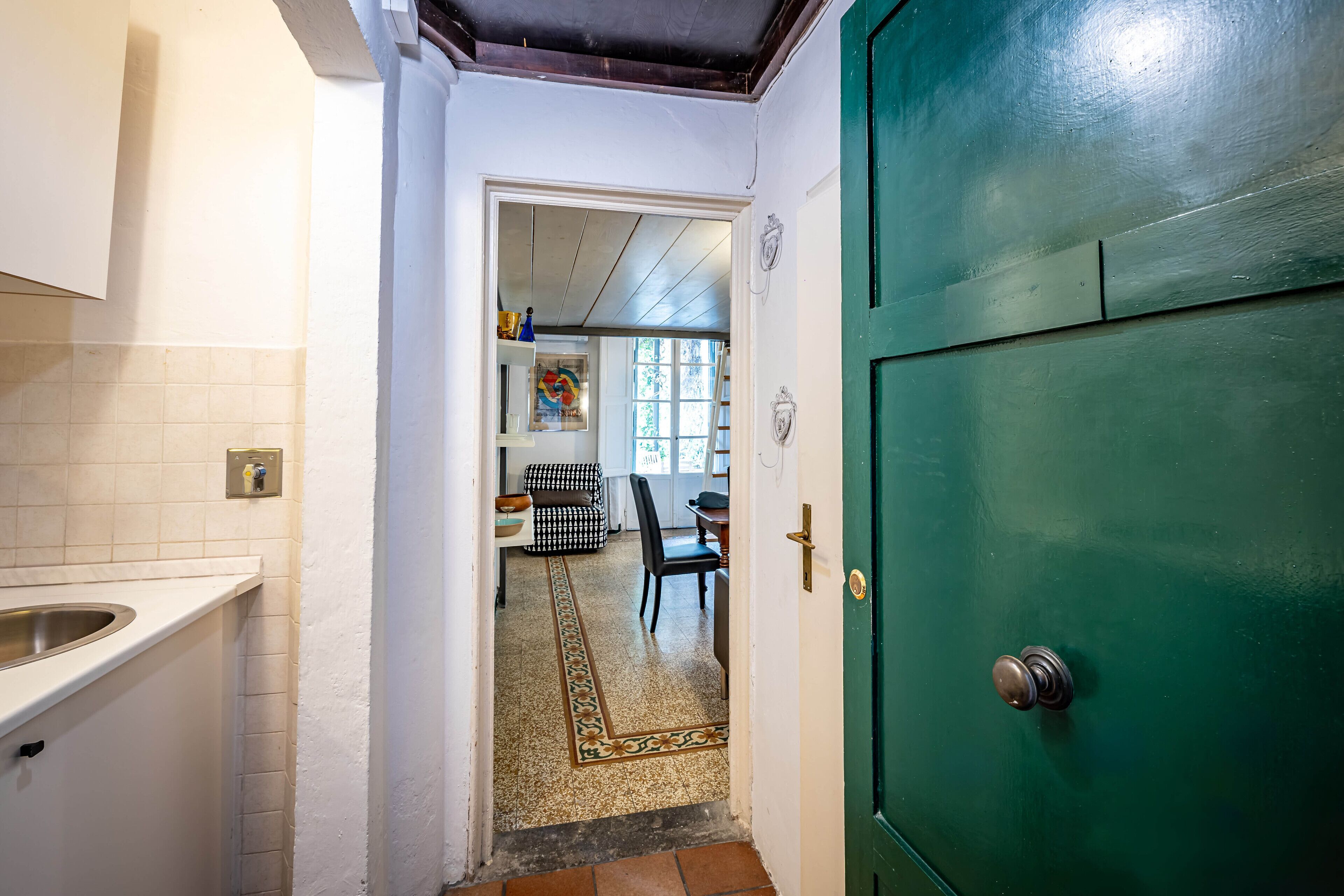 A narrow hallway with a green door, a tiled floor, and a view into a dining area with a patterned rug.