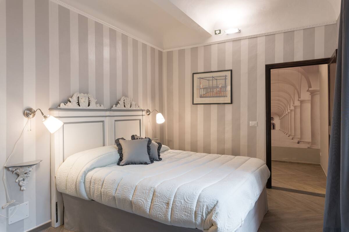 Une chambre à coucher avec un lit, des lampes de chevet, un tableau encadré et une vue sur un couloir.