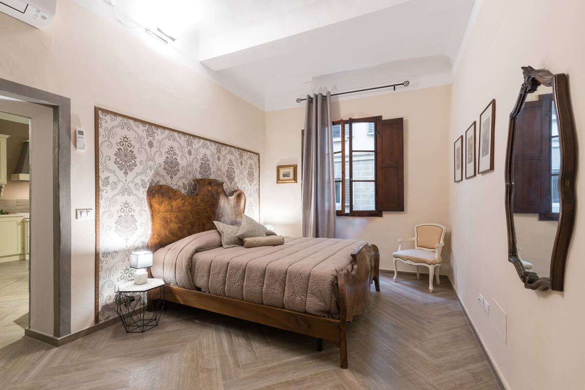 Une chambre à coucher avec un lit en bois, une table de chevet, une chaise, un grand miroir et une fenêtre avec des rideaux.
