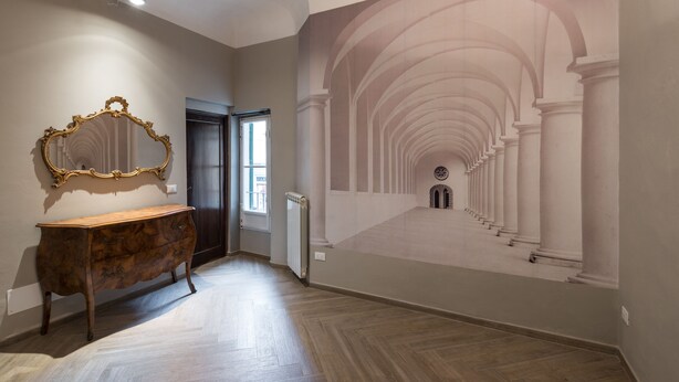 A hallway with a wooden floor, a large ornate mirror, and a mural of an arched corridor. - Flospirit - San Lorenzo (Florence)