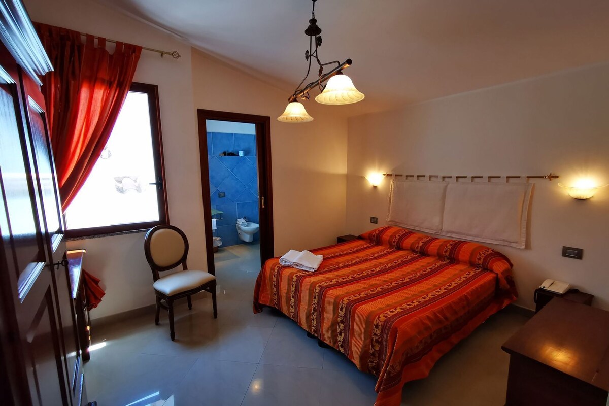 A bedroom with a bed, a chair, a desk, a window with curtains, and a bathroom visible through an open door.