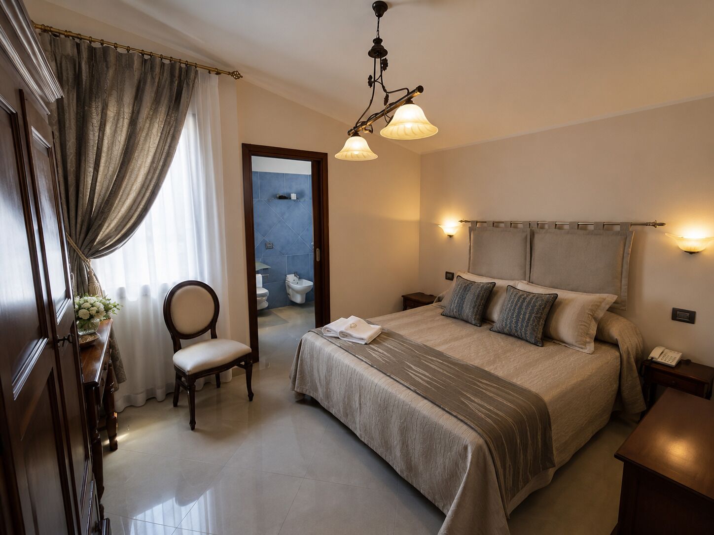 A hotel room with a bed, a chair, a bedside table, a nightstand, a mirror, a chandelier, and a bathroom visible through an open door.