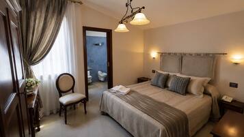 A hotel room with a bed, a chair, a bedside table, a nightstand, a mirror, a chandelier, and a bathroom visible through an open door.