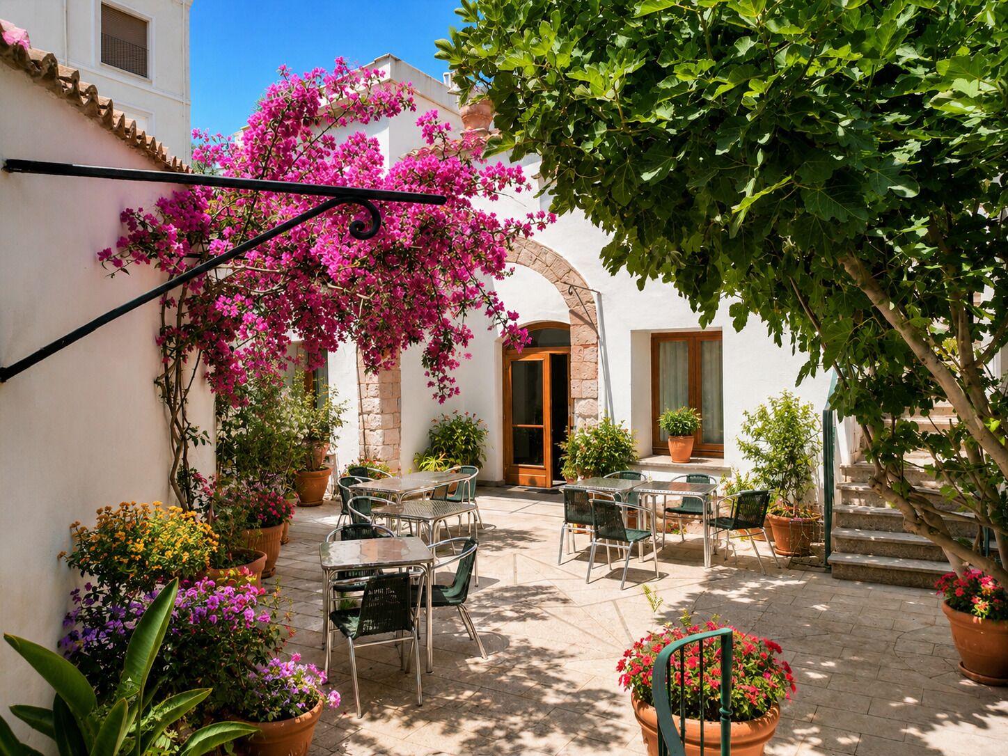 Une terrasse extérieure avec des tables et des chaises, entourée de plantes en pot et d’arbustes en fleurs.