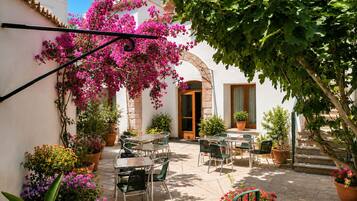 Une terrasse extérieure avec des tables et des chaises, entourée de plantes en pot et d’arbustes en fleurs.