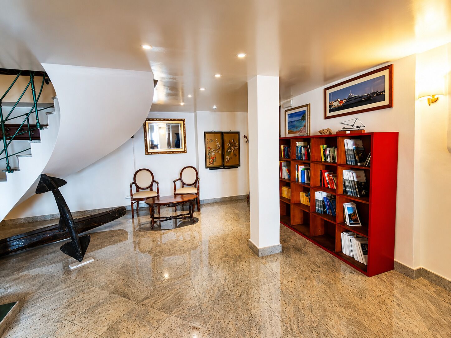 Un pasillo amplio con suelo de mármol, una estantería roja, dos sillas, una escalera y obras enmarcadas en las paredes.