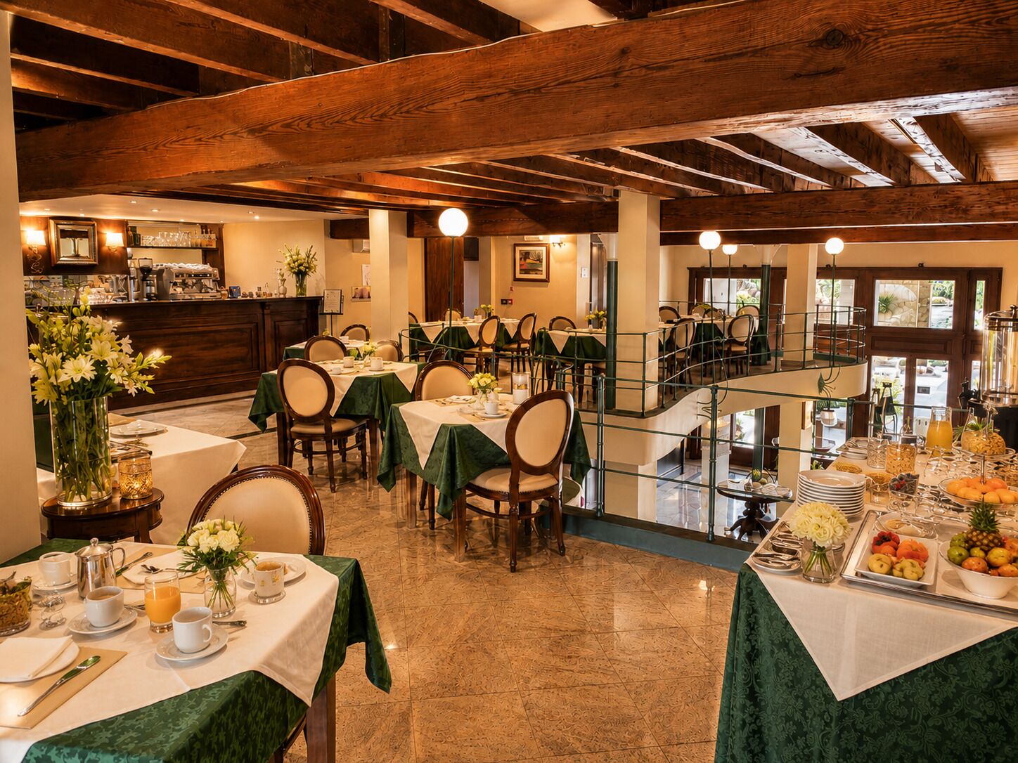 A dining area with tables set for breakfast, a buffet with fruits and pastries, and a bar area with coffee machines.