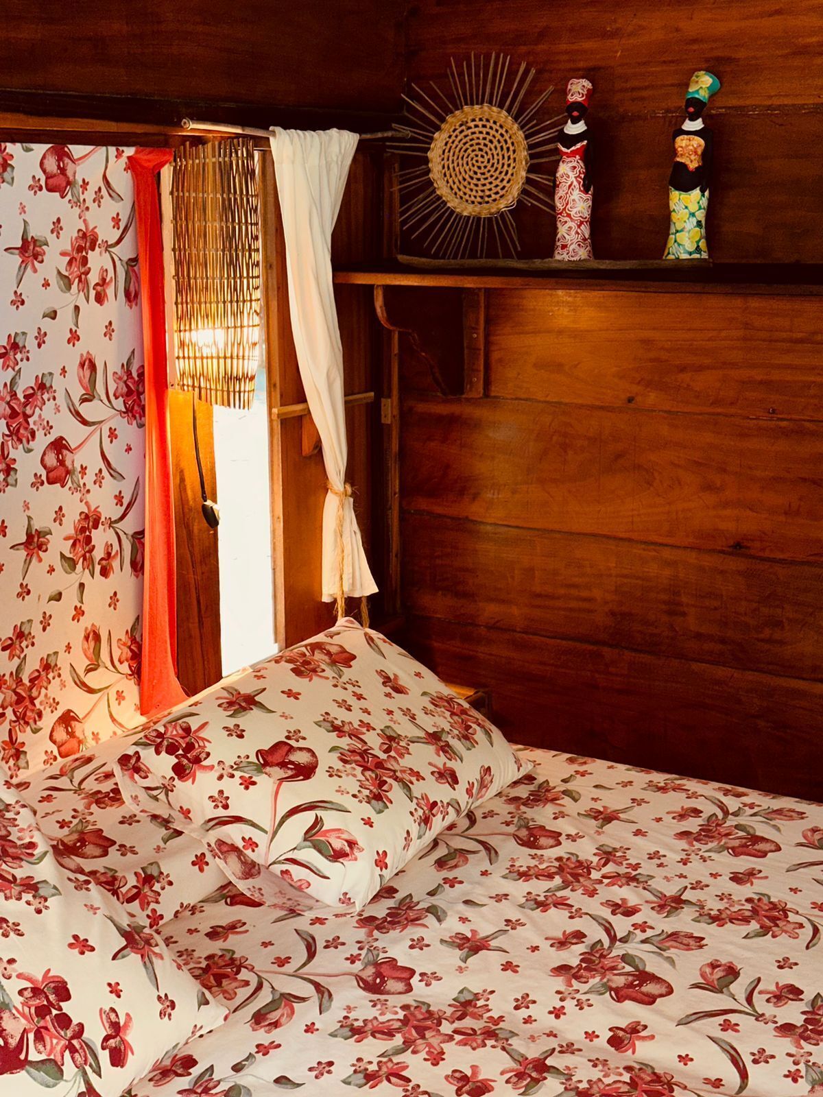 A wooden room with a bed, a window with floral curtains, and a shelf with decorative items.