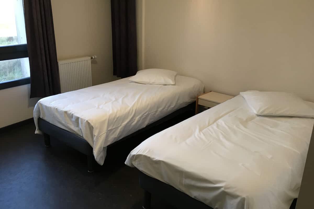 A room with two single beds, a small bedside table, a window with curtains, and a radiator.