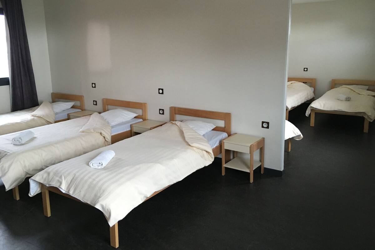 A hotel room with four single beds, each with a wooden headboard, a bedside table, and a rolled towel on the bed.