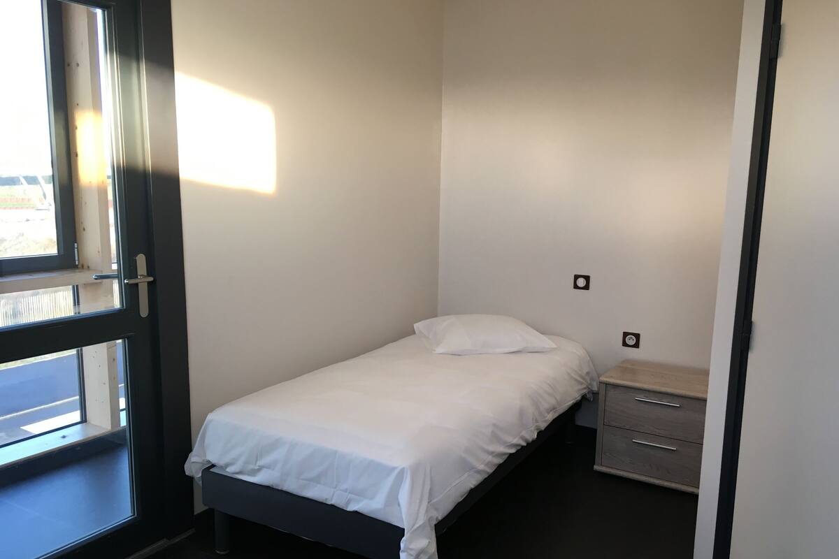 A small, neatly made bed in a room with a window, a bedside table, and a wall socket.