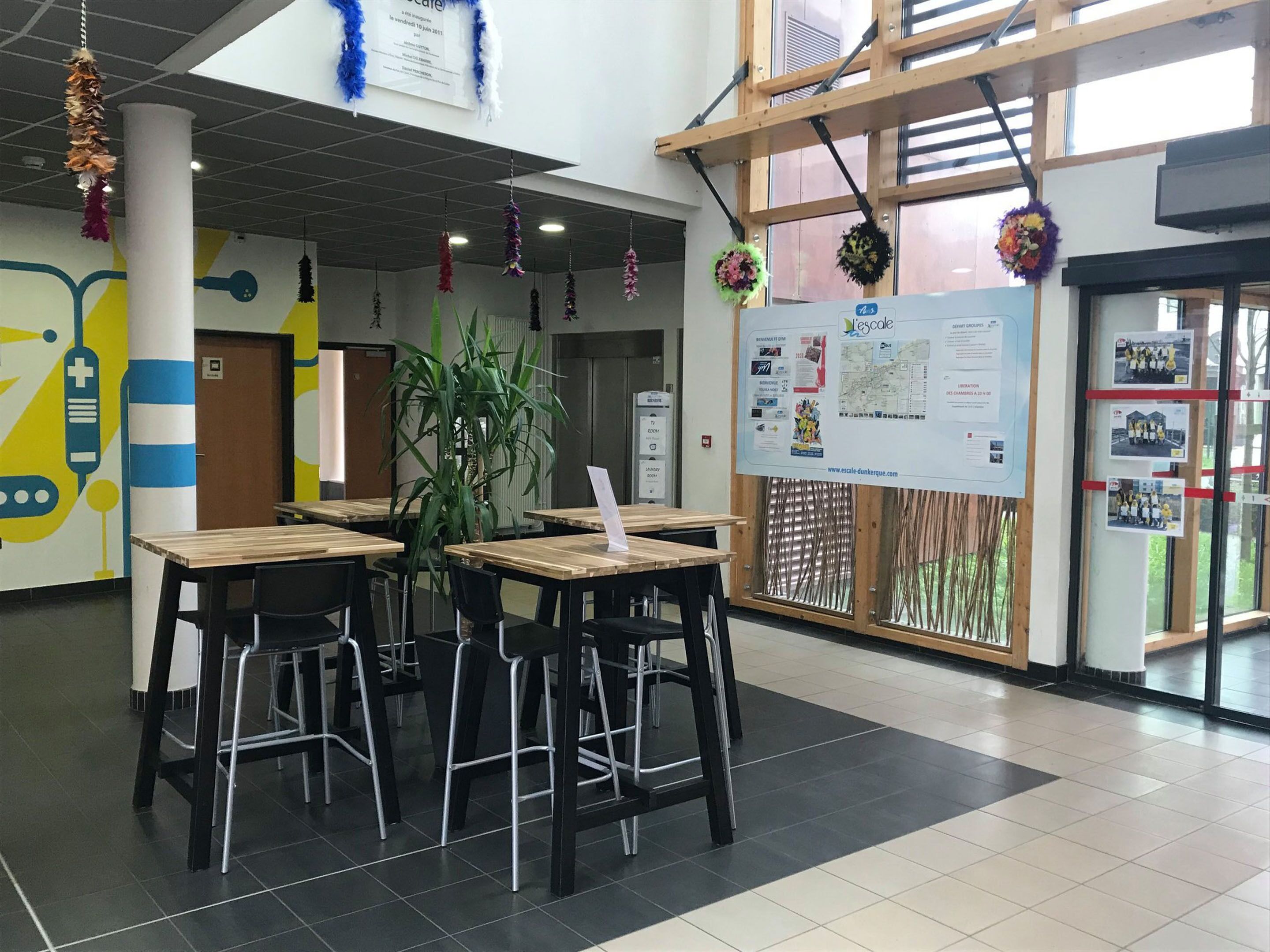Una cafetería con mesas y sillas, un cartel grande en la pared y una planta en el centro.
