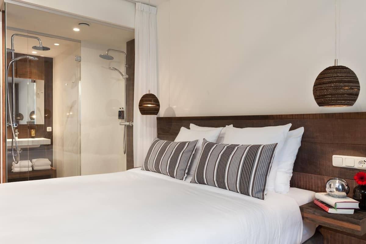 Une chambre d’hôtel moderne avec un grand lit, une tête de lit en bois, une table de chevet avec un livre et un vase, et une douche séparée par des parois en verre.