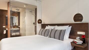Una habitación de hotel moderna con una cama grande, cabecera de madera, una mesita de noche con un libro y un jarrón, y una zona de ducha con mampara de cristal.
