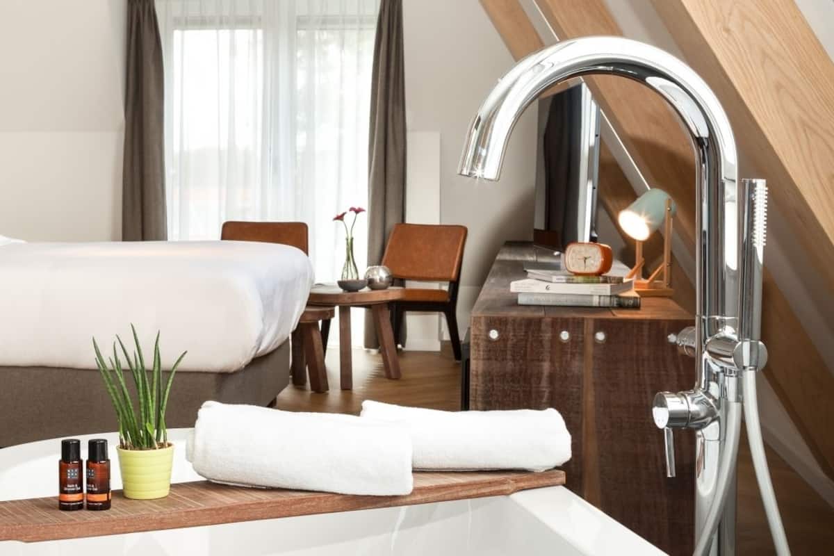 Une chambre d’hôtel moderne dotée d’un grand lit, d’une table de chevet en bois avec des articles de toilette, d’un petit bureau avec une lampe et d’une salle de bain avec une douche moderne.
