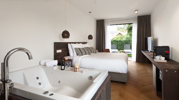 A modern hotel room with a large bathtub, a neatly made bed, a desk with a coffee machine, and a view of the outdoors. - Van Heeckeren Hotel (Nes)