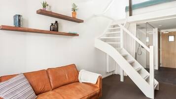 A modern living room with a brown leather sofa, white walls, and wooden shelves with decorative items. A white spiral staircase leads to an upper level.