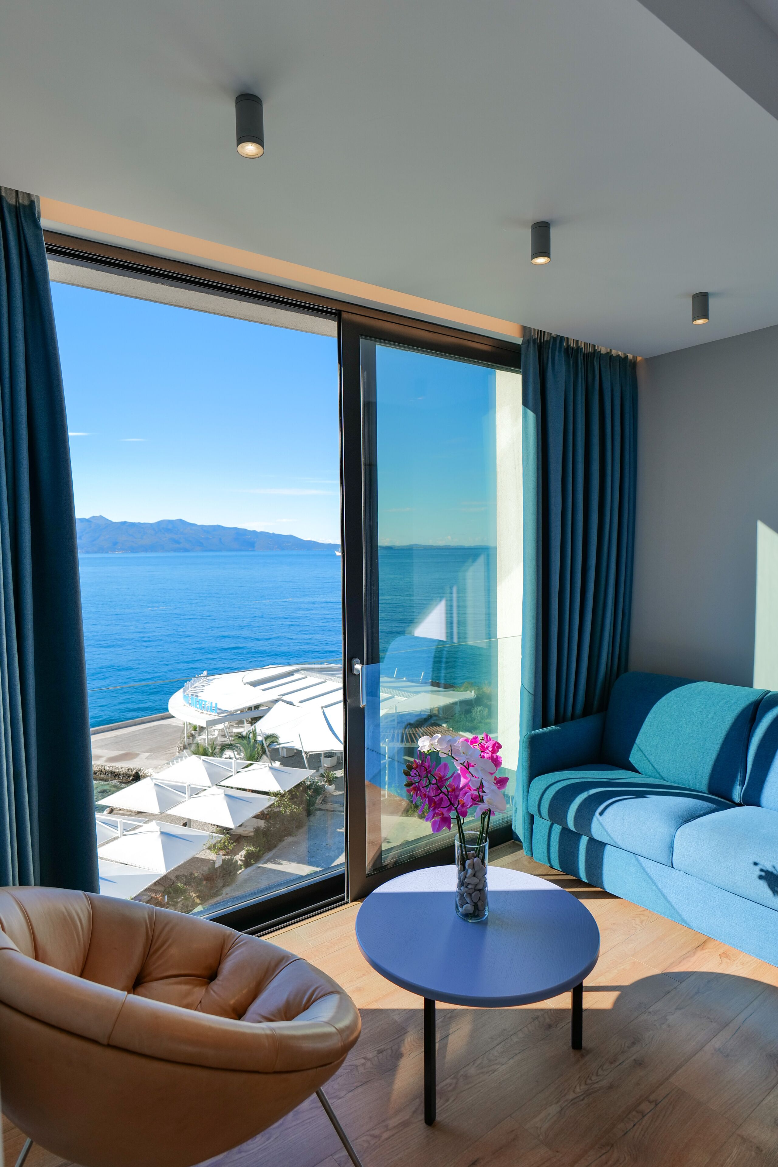 Un salon moderne avec vue sur la mer, comprenant un canapé, un fauteuil et une petite table agrémentée de fleurs.