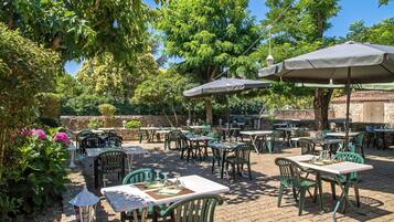 Een terras buiten met tafels en stoelen, omringd door groen en bomen.
