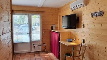 Una habitación pequeña con paredes de madera, un televisor montado en la pared, una mesita con una silla y una ventana con persianas.