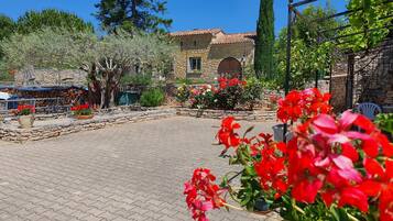 Un patio con pavimento de piedra, un edificio de piedra con techo de tejas y diversas macetas con plantas y flores.