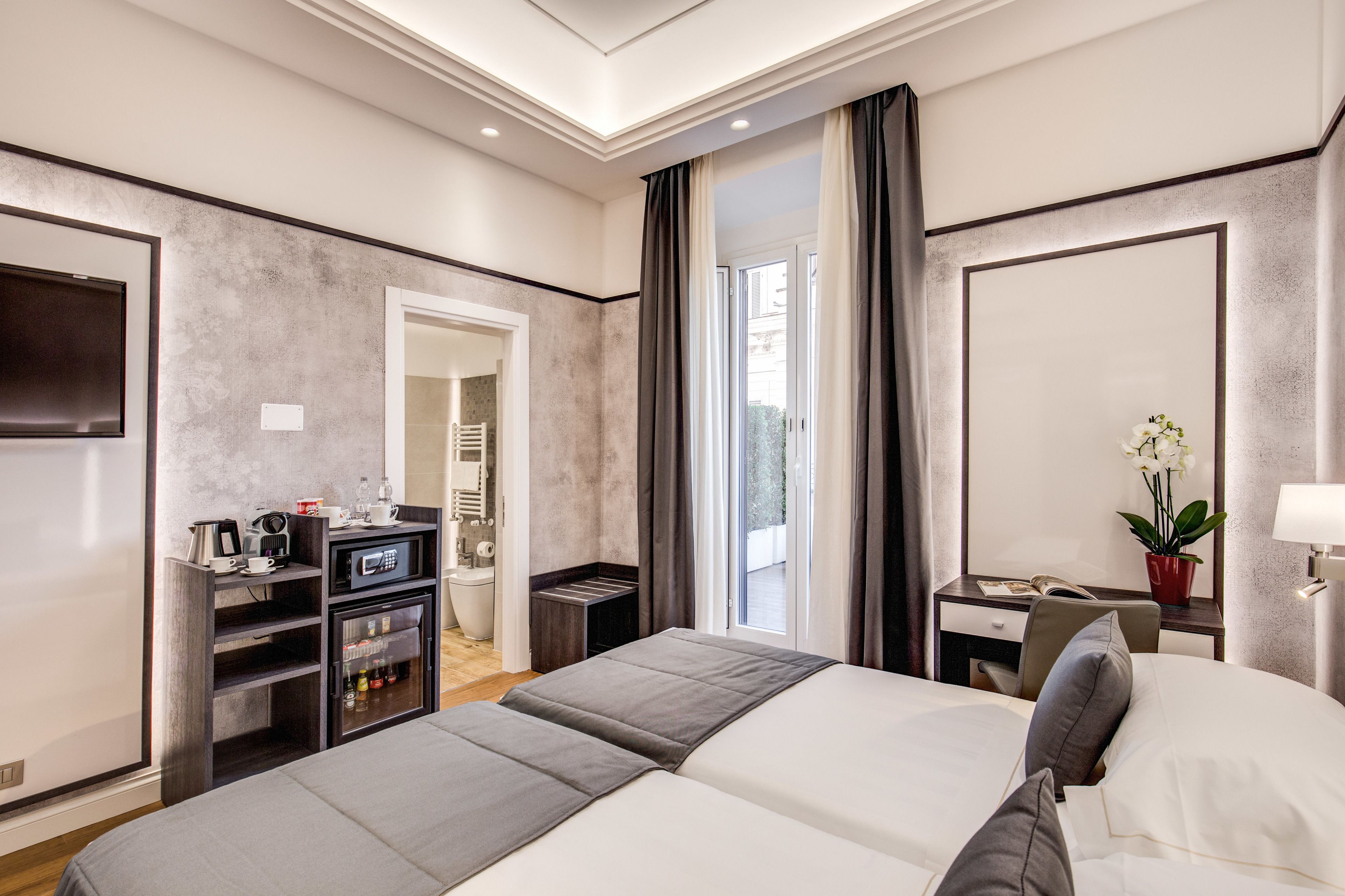 A modern hotel room with a large bed, a desk, a chair, a small kitchenette, and a bathroom visible through an open door.