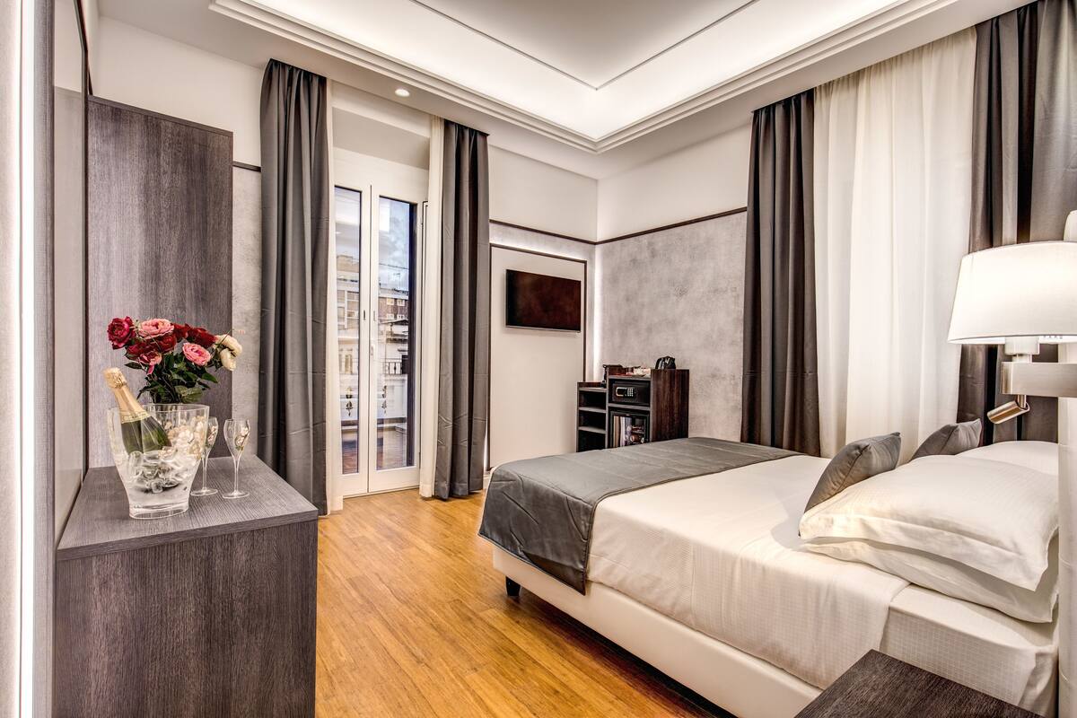 A modern hotel room with a large bed, a bedside table with a vase of flowers, a flat-screen TV, and a view of the city through the window.