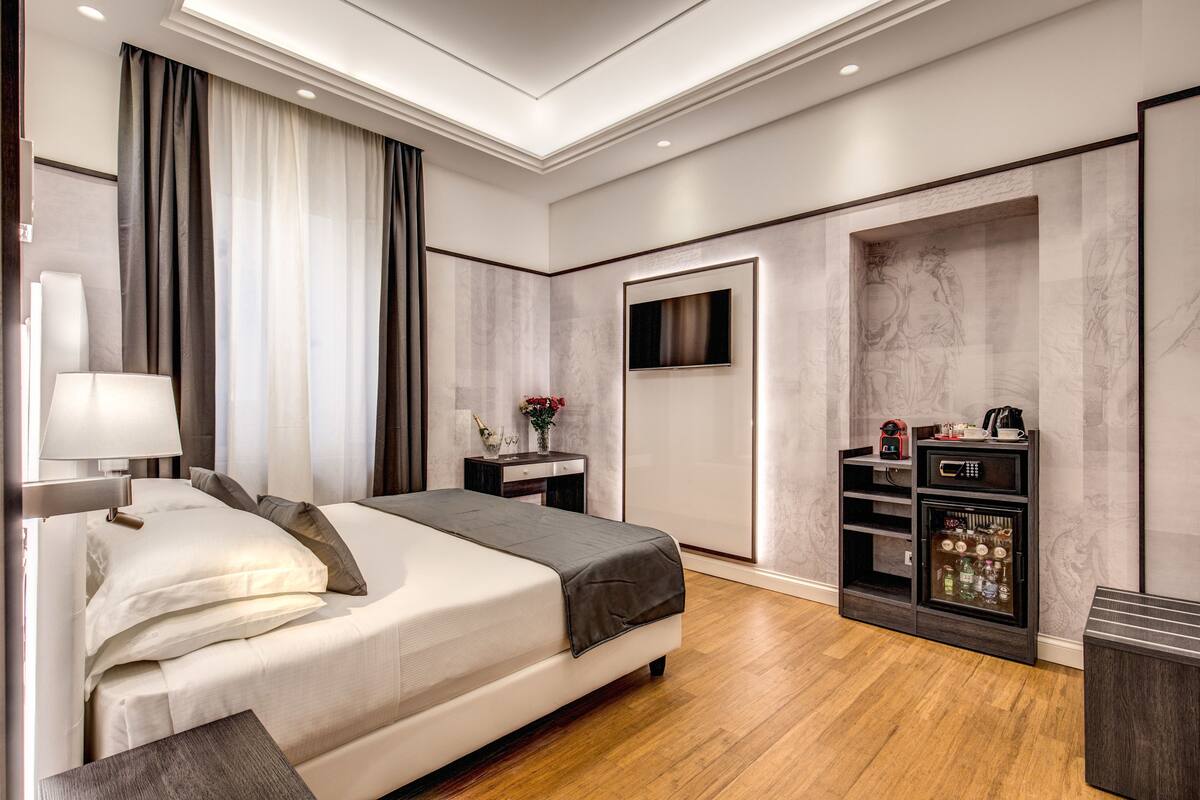 A modern hotel room with a large bed, a bedside table, a TV mounted on the wall, and a minibar.