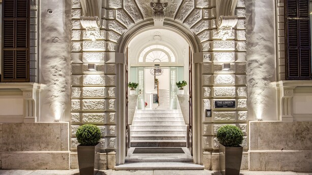 Ein beeindruckender Eingangsbereich mit einer Marmortreppe, gesäumt von Säulen und Pflanzen in Töpfen, der zu einer Tür mit dem Schild "The Liberty Boutique Hotel" führt.