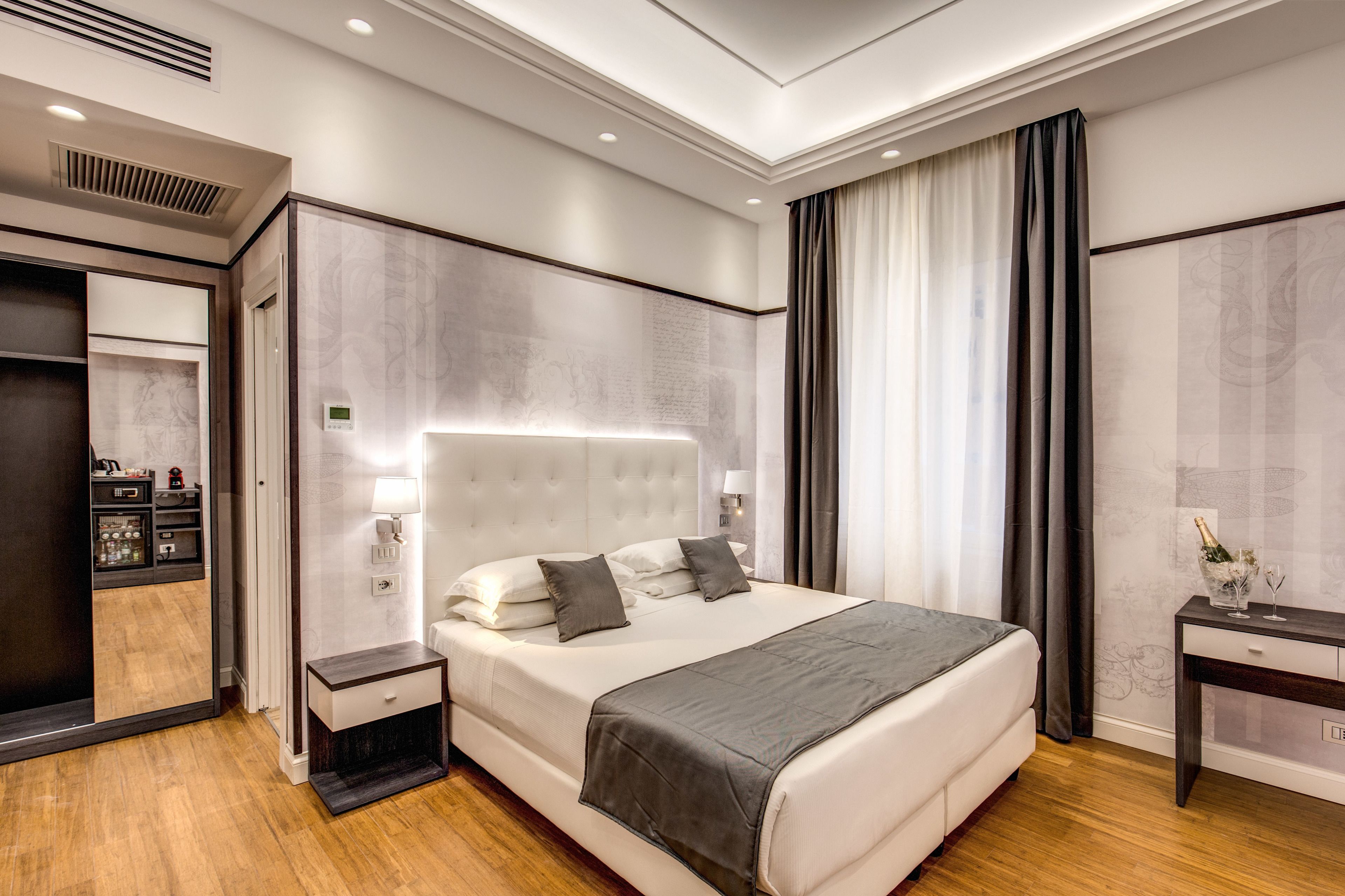 A modern hotel room with a large bed, bedside tables, a wardrobe, and a desk with a lamp.