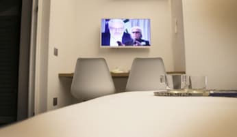 Habitación de hotel con televisión montada en la pared, dos sillones y una mesa con un vaso y un libro.